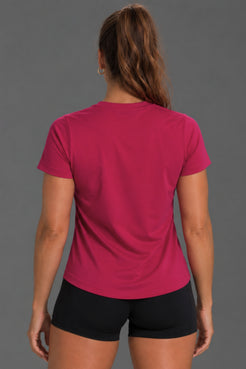 A woman viewed from the back wearing a bright pink short-sleeve athletic T-shirt and black shorts against a solid gray studio background. || Berry