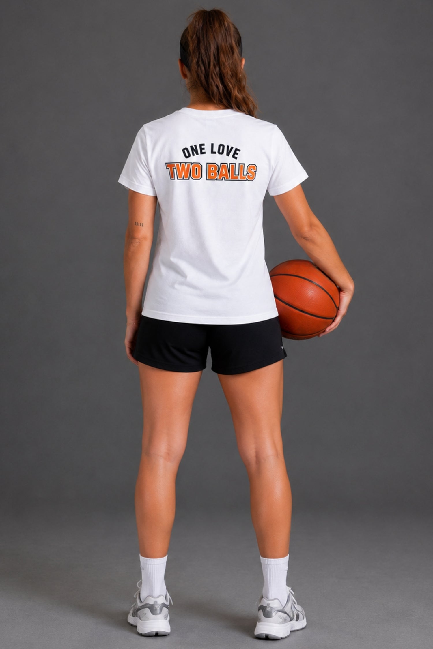 A woman stands with her back to the camera wearing a white graphic t-shirt with 'ONE LOVE TWO BALLS' printed on the back, black athletic shorts, white socks, and gray sneakers, while holding a basketball against a solid gray studio background.