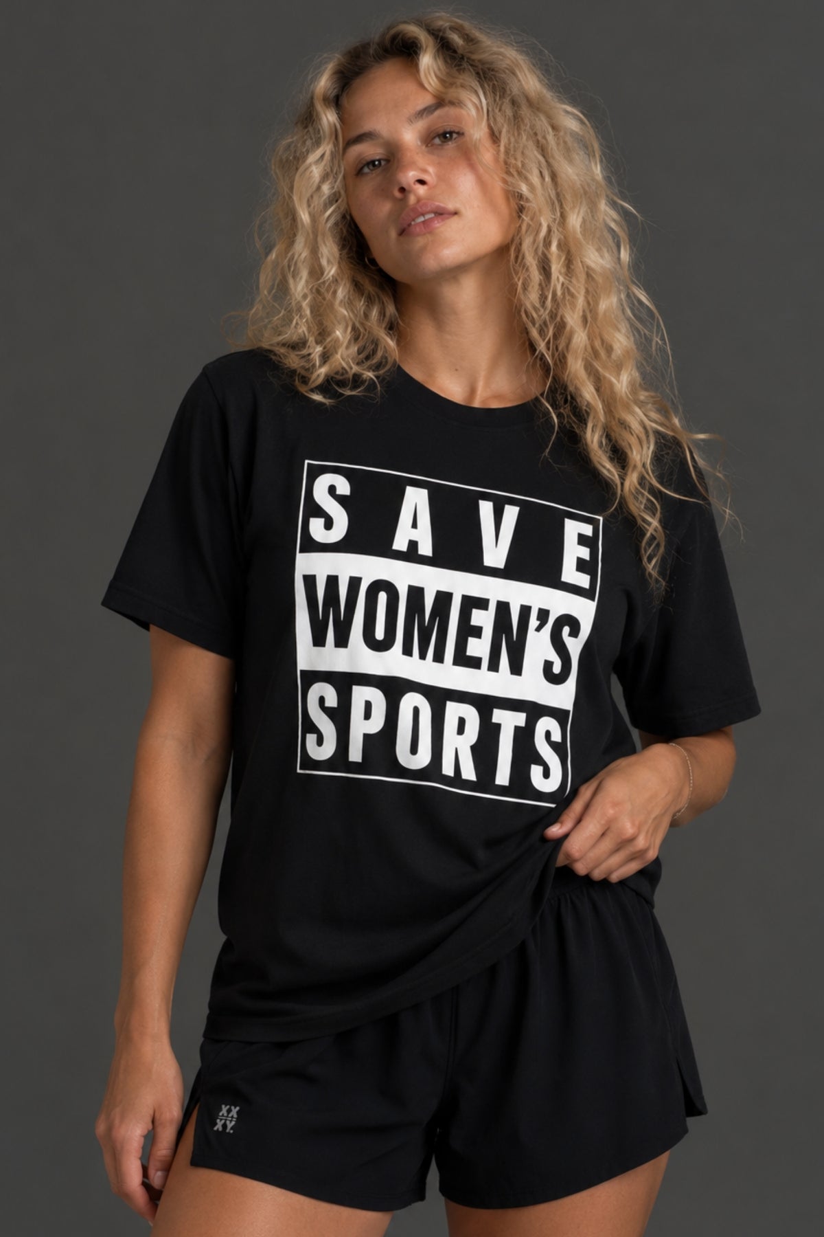 A woman wearing a black graphic t-shirt with the text 'SAVE WOMEN'S SPORTS' in white block letters, paired with black athletic shorts, against a dark gray studio background. || Black