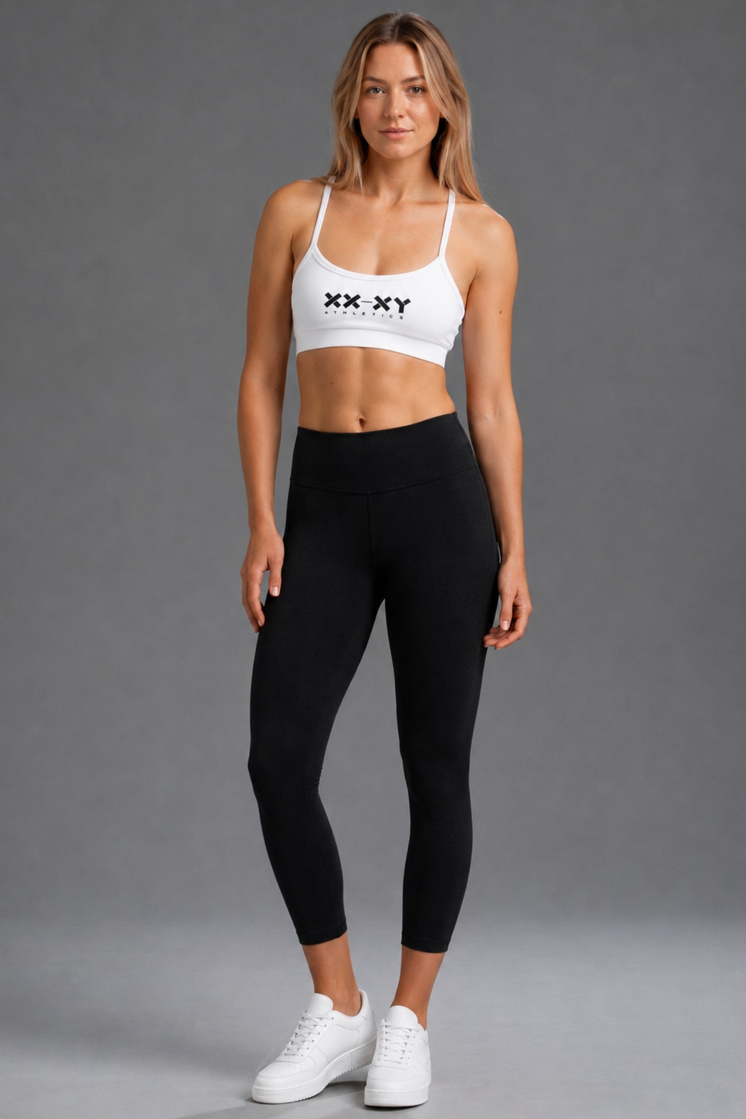 A woman modeling a white sports bra with 'XX XY' printed on the chest and black high-waisted leggings, standing against a solid gray studio background. || Black Heather