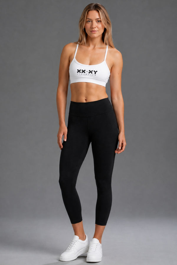 A woman modeling a white sports bra with 'XX XY' printed on the chest and black high-waisted leggings, standing against a solid gray studio background. || Black Heather