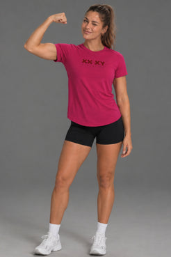 A woman posing in a bright pink short-sleeve athletic t-shirt with a black 'XX XY' logo on the chest, paired with black athletic shorts, white socks, and white sneakers, against a solid gray studio background. || Berry