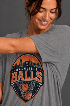 A woman wearing a heather gray t-shirt with a large Nashville Balls graphic on the front, featuring orange and navy blue basketball-themed cityscape artwork. || Dark Heather Gray