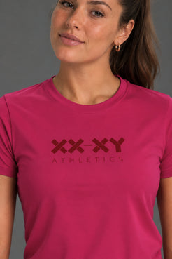 A woman wearing a bright pink crewneck athletic t-shirt with a dark red 'XX-XY ATHLETICS' logo printed on the chest, set against a solid gray background. || Berry
