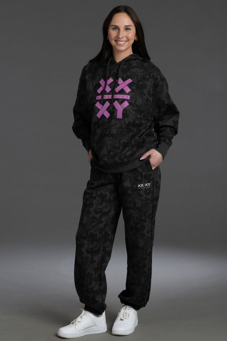Person wearing a black hoodie and pants with pink logo on a gray background || Black Camo
