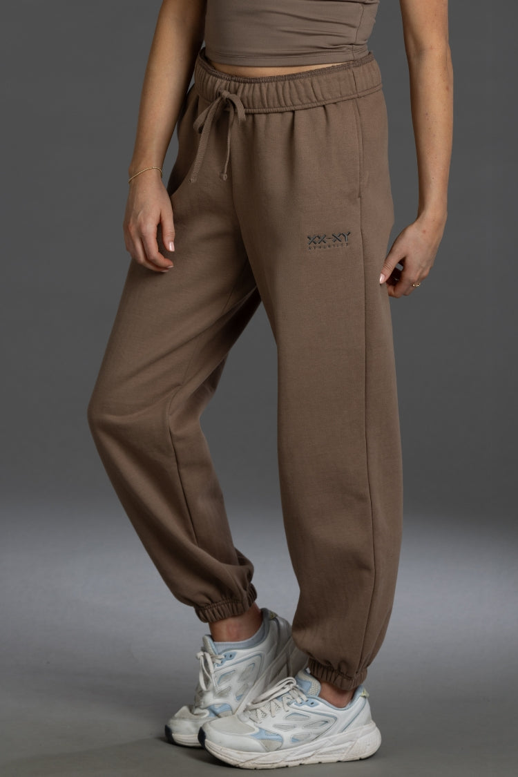 A person wearing brown jogger sweatpants with an elastic drawstring waist and elastic cuffs, paired with white sneakers, shown from the waist down against a dark gray studio background. || Mocha