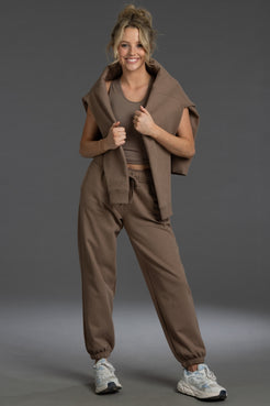A woman modeling a coordinated taupe-brown activewear set consisting of a sleeveless cropped vest, a cropped tank top, jogger-style pants, and a draped shawl over her shoulders, paired with white sneakers against a dark gray studio background. || Mocha
