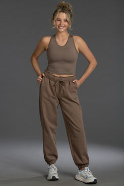 A woman modeling a matching brown activewear set consisting of a cropped tank top and jogger sweatpants, paired with white sneakers, against a solid gray studio background. || Mocha