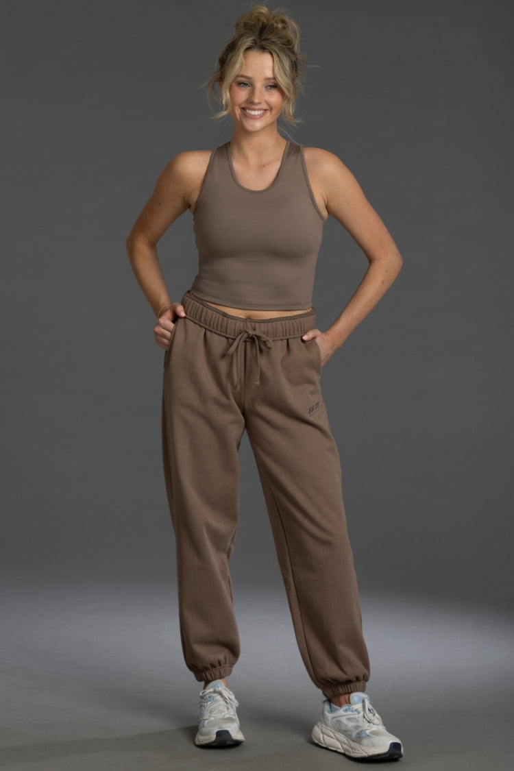 A woman modeling a matching brown activewear set consisting of a cropped tank top and jogger sweatpants, paired with white sneakers, against a solid gray studio background. || Mocha