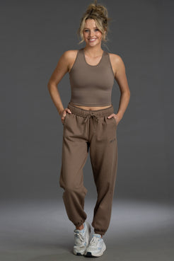 A woman modeling a matching taupe-brown activewear set consisting of a ribbed tank top and jogger sweatpants, paired with white sneakers, against a solid gray studio background. || Mocha