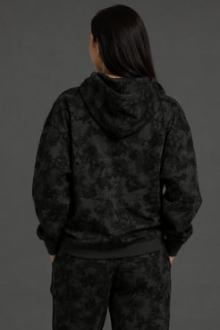 A person shown from the back wearing a black hooded sweatshirt and matching black patterned pants against a dark gray studio background. || Black Camo