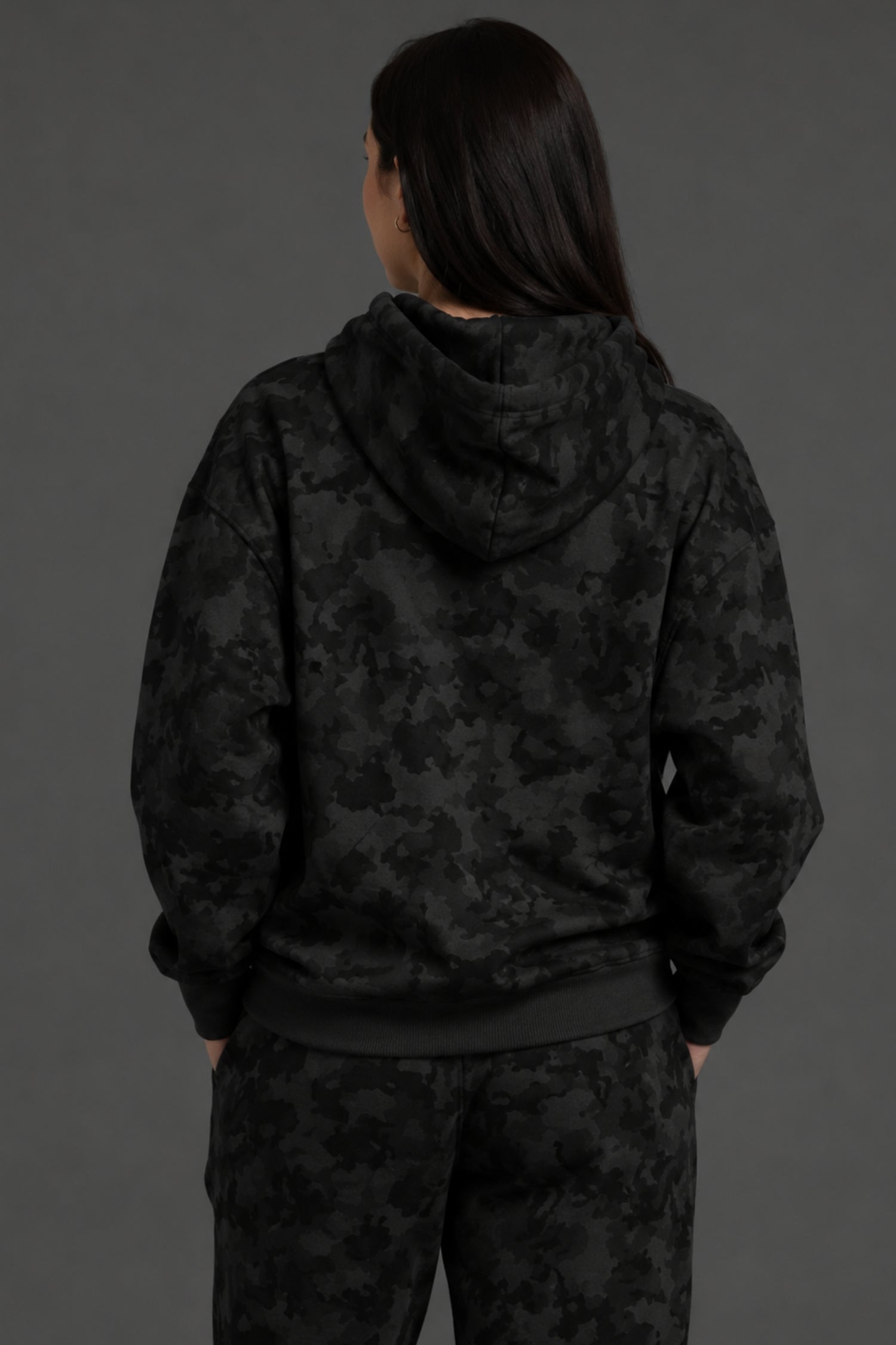 A person shown from the back wearing a black hooded sweatshirt and matching black patterned pants against a dark gray studio background. || Black Camo