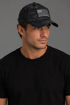 A man wearing a black camouflage baseball cap with a black and white American flag patch and 'ROOT ATHLETICS' text, paired with a black t-shirt against a solid gray background. || Black Camo