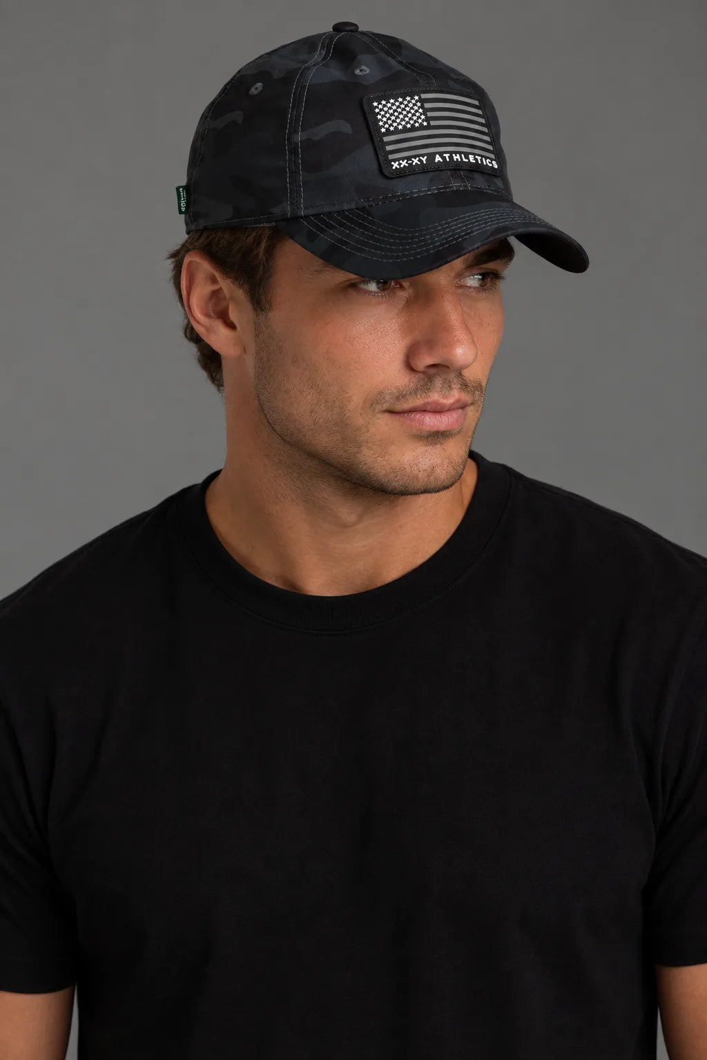 A man wearing a black camouflage baseball cap with a black and white American flag patch and 'ROOT ATHLETICS' text, paired with a black t-shirt against a solid gray background. || Black Camo