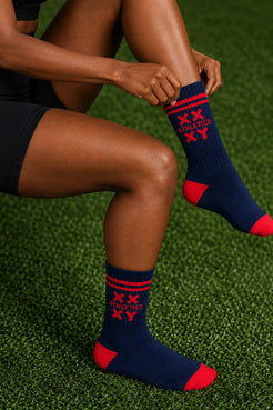 Black female model wearing navy crew socks with red stripes, red toe and heel accents, and a red XX-XY Athletics logo while adjusting the sock on green turf. || blue aster/red