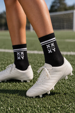 Female athlete wearing black XX-XY Athletics crew socks with white stripes and white soccer cleats on a soccer field. || Black