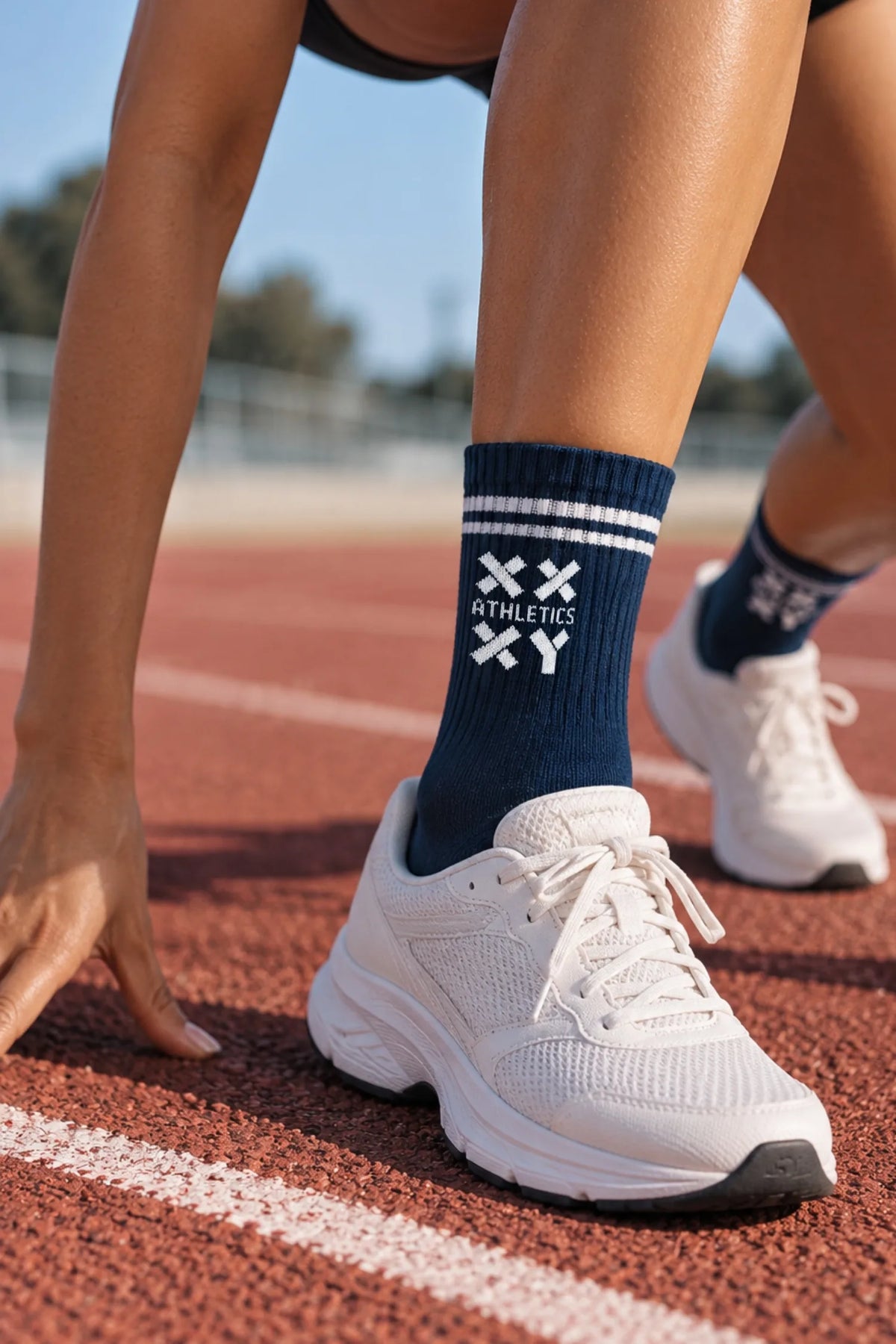 Female runner in a starting position on an outdoor track, wearing navy XX-XY Athletics crew socks with white stripes and white running shoes. || Navy