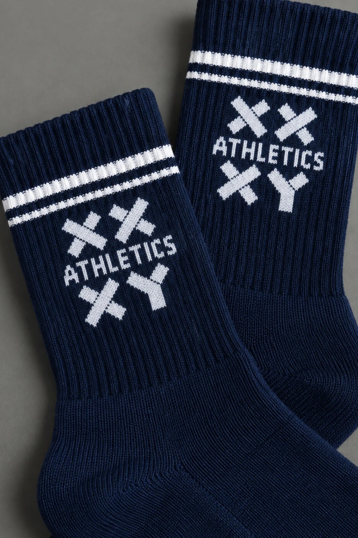 Close-up of navy XX-XY Athletics crew socks featuring white stripes and a white knit logo on a gray background. || Navy