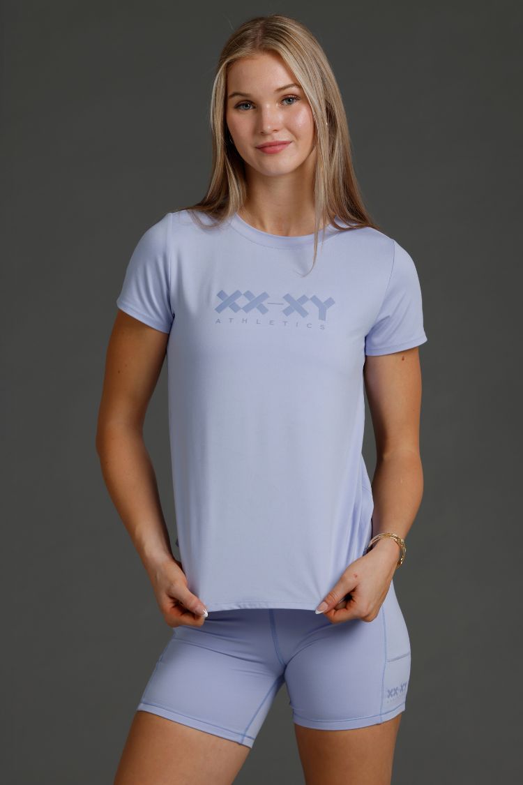 Woman wearing a Lavender t-shirt and shorts with a logo on a gray background || Lavender Gray