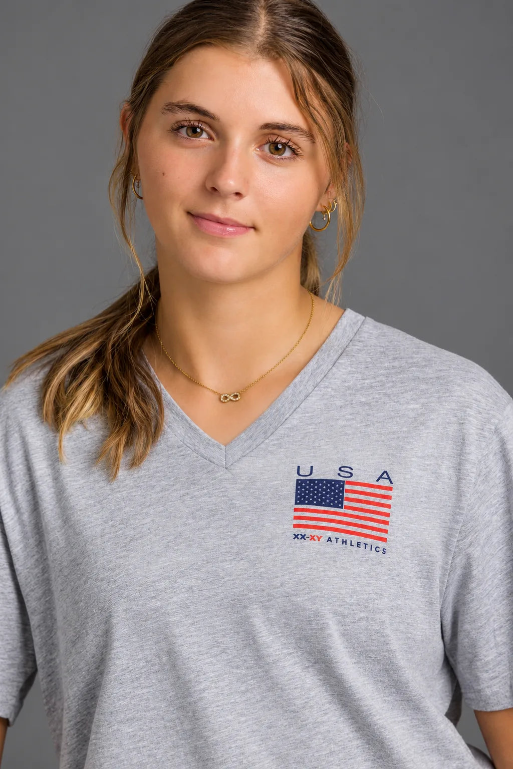 Female softball player wearing a gray USA XX-XY Athletics V-neck T-shirt against a gray studio background. || Light Heather Gray