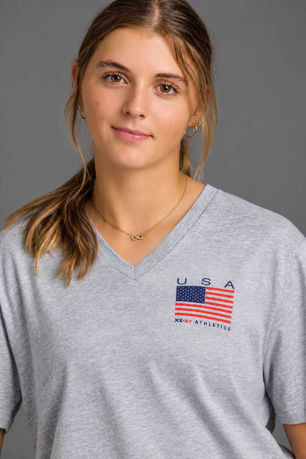 Female softball player wearing a gray USA XX-XY Athletics V-neck T-shirt against a gray studio background. || Light Heather Gray