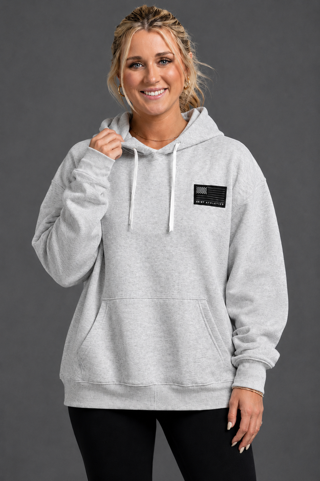 Woman wearing a gray hoodie with a small patch on a dark background || Light Heather Gray