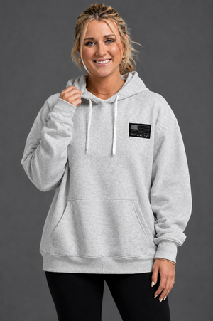Woman wearing a gray hoodie with a small patch on a dark background || Light Heather Gray
