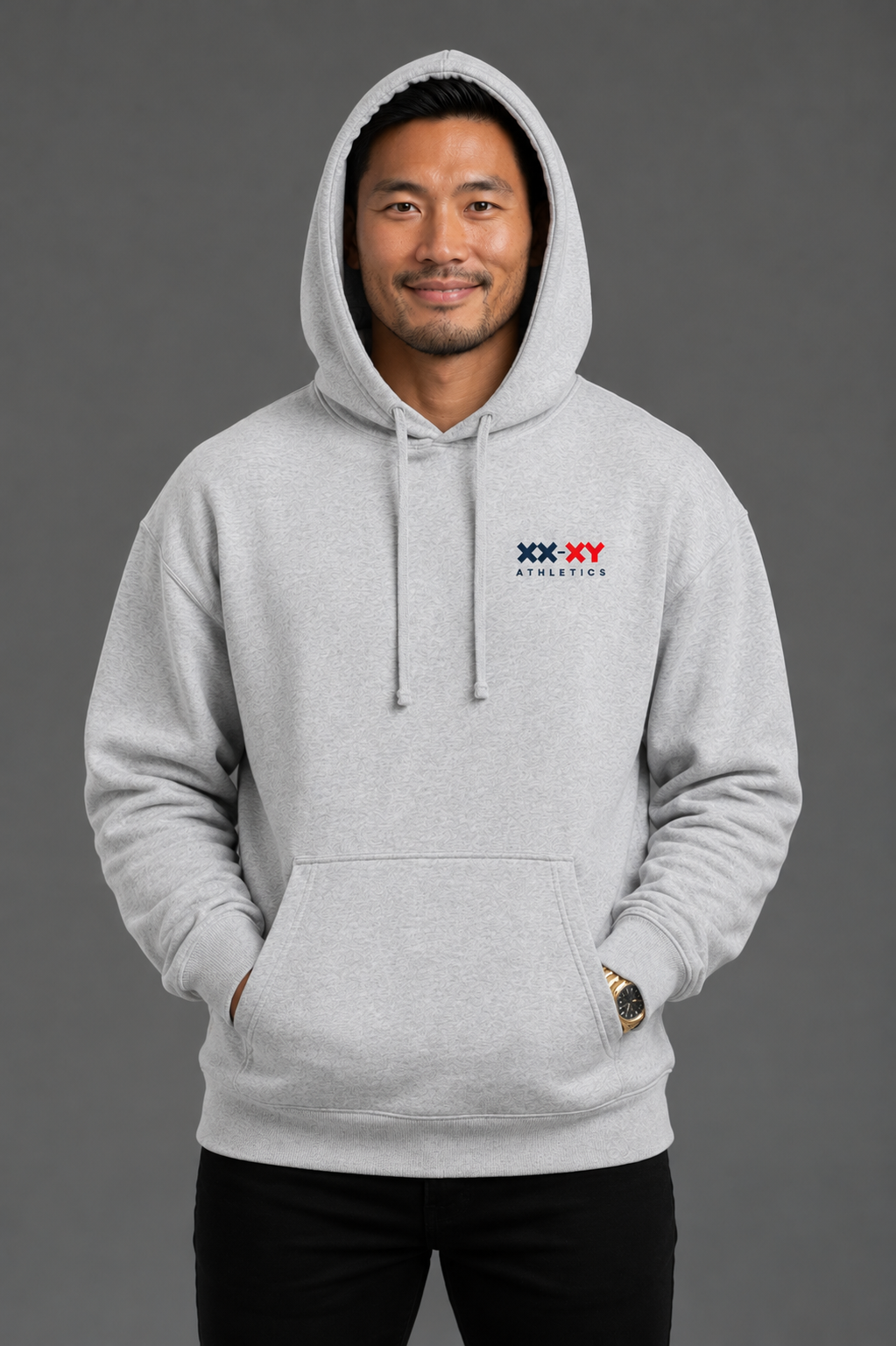 A man wearing a light gray heather pullover hoodie with a small red, white, and blue 'XX-XY Athletics' logo on the left chest, set against a solid dark gray studio background. || Heather Gray
