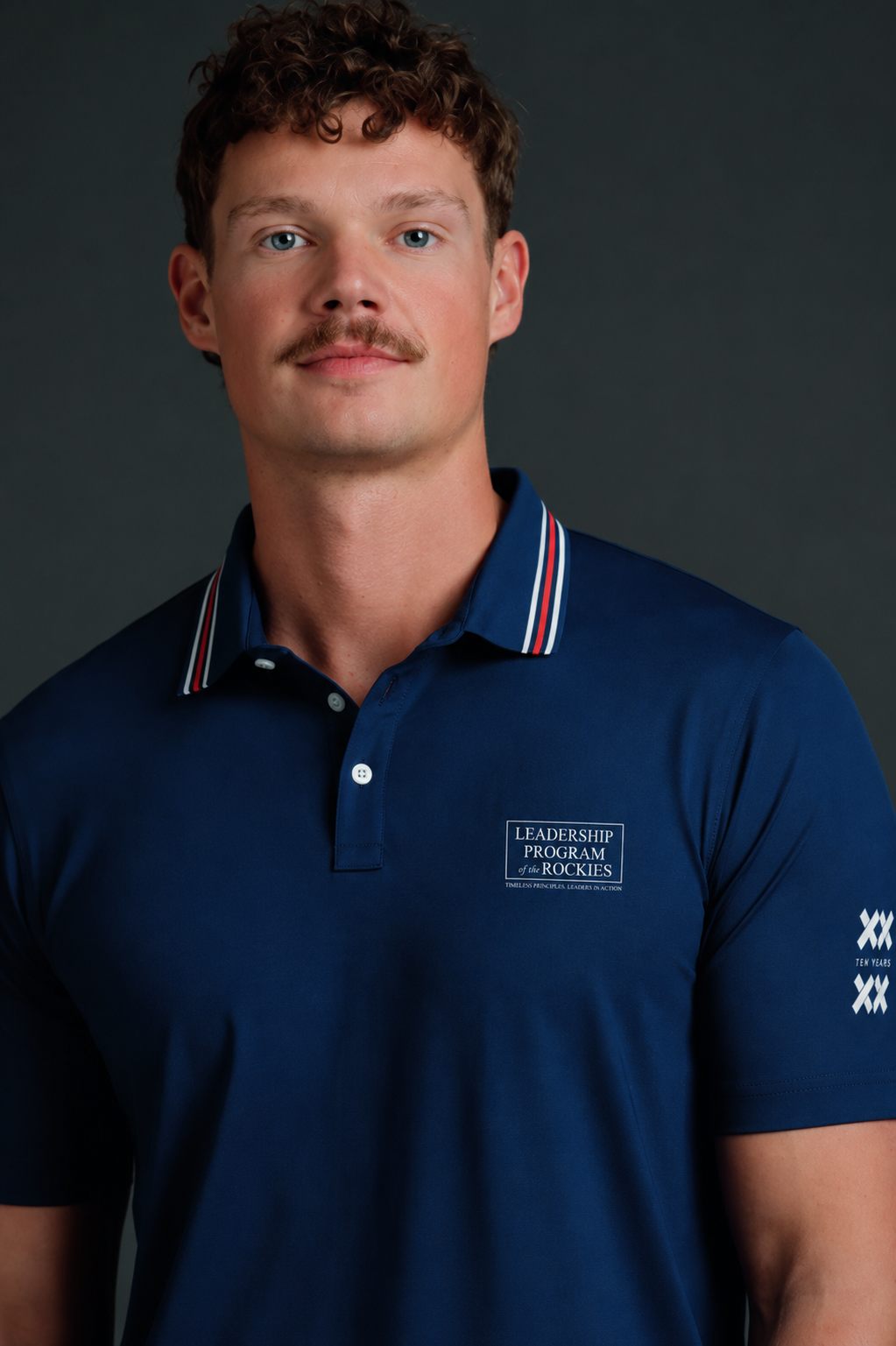 Man wearing a navy blue polo shirt with logos on a dark background