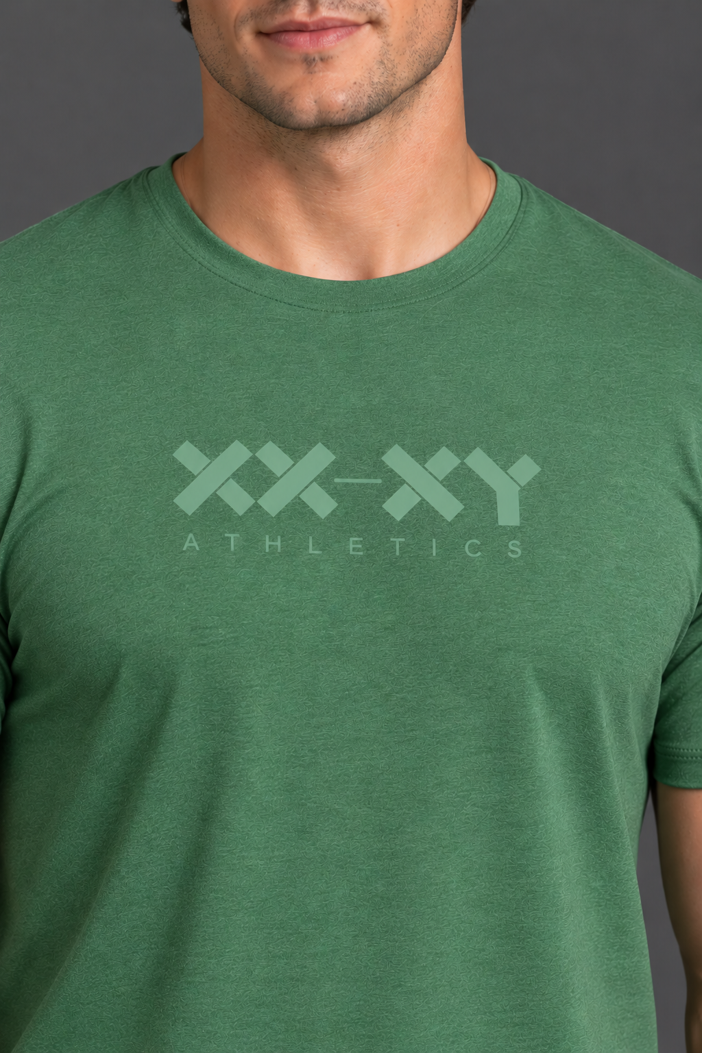 A close-up of a man wearing a heather green crewneck t-shirt with a light green 'XX-XY ATHLETICS' logo printed on the chest, set against a dark gray background. || Hunter Green Heather