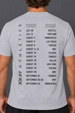 A man wearing a white t-shirt, viewed from the back. The shirt features a black printed schedule of '2026 Tour' dates for 'Athletics' on the back. || Light Heather Gray