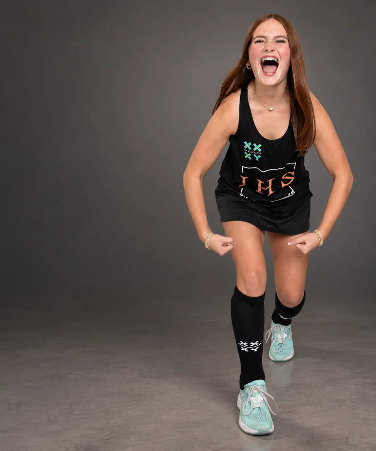 A young woman in a black athletic tank top, black skirt, black knee-high socks, and mint green sneakers poses dynamically against a dark gray studio backdrop. The tank top features a graphic with the letters 'IHS' in orange and teal.