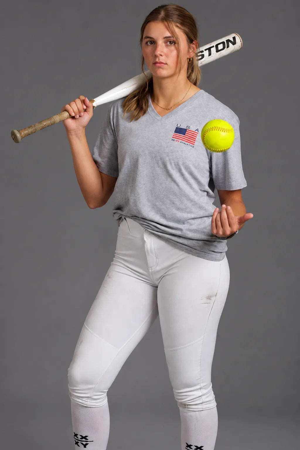 Female softball player wearing a gray USA XX-XY Athletics V-neck T-shirt, holding a softball and bat. || Light Heather Gray