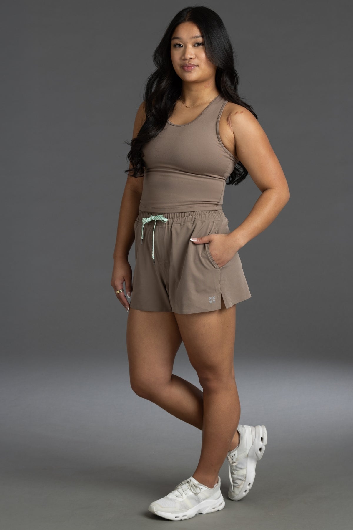 A woman modeling a matching taupe-brown activewear set consisting of a ribbed tank top and shorts with a mint green drawstring, paired with white sneakers, against a solid gray studio background. || Mocha