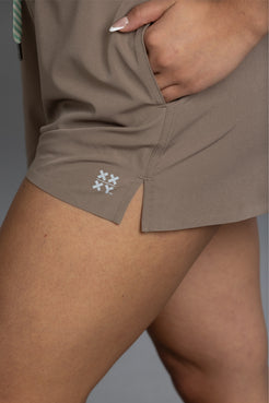 Close-up of a person wearing taupe-brown athletic shorts with a side pocket and a small white embroidered snowflake logo near the hem. || Mocha