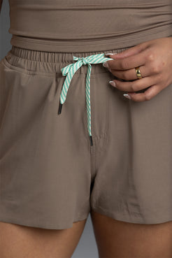 Close-up of a person wearing taupe-brown shorts with an elastic waistband and a mint green and white striped drawstring tied at the front. A hand with a gold ring is adjusting the drawstring. || Mocha