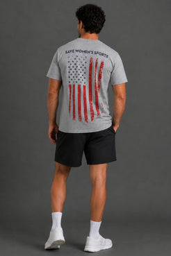 A man viewed from behind wearing a heather gray t-shirt with a distressed American flag graphic and the text 'SAVES WOMEN'S SPORTS' on the back, paired with black athletic shorts, white socks, and white sneakers against a dark gray studio background. || Heather Gray