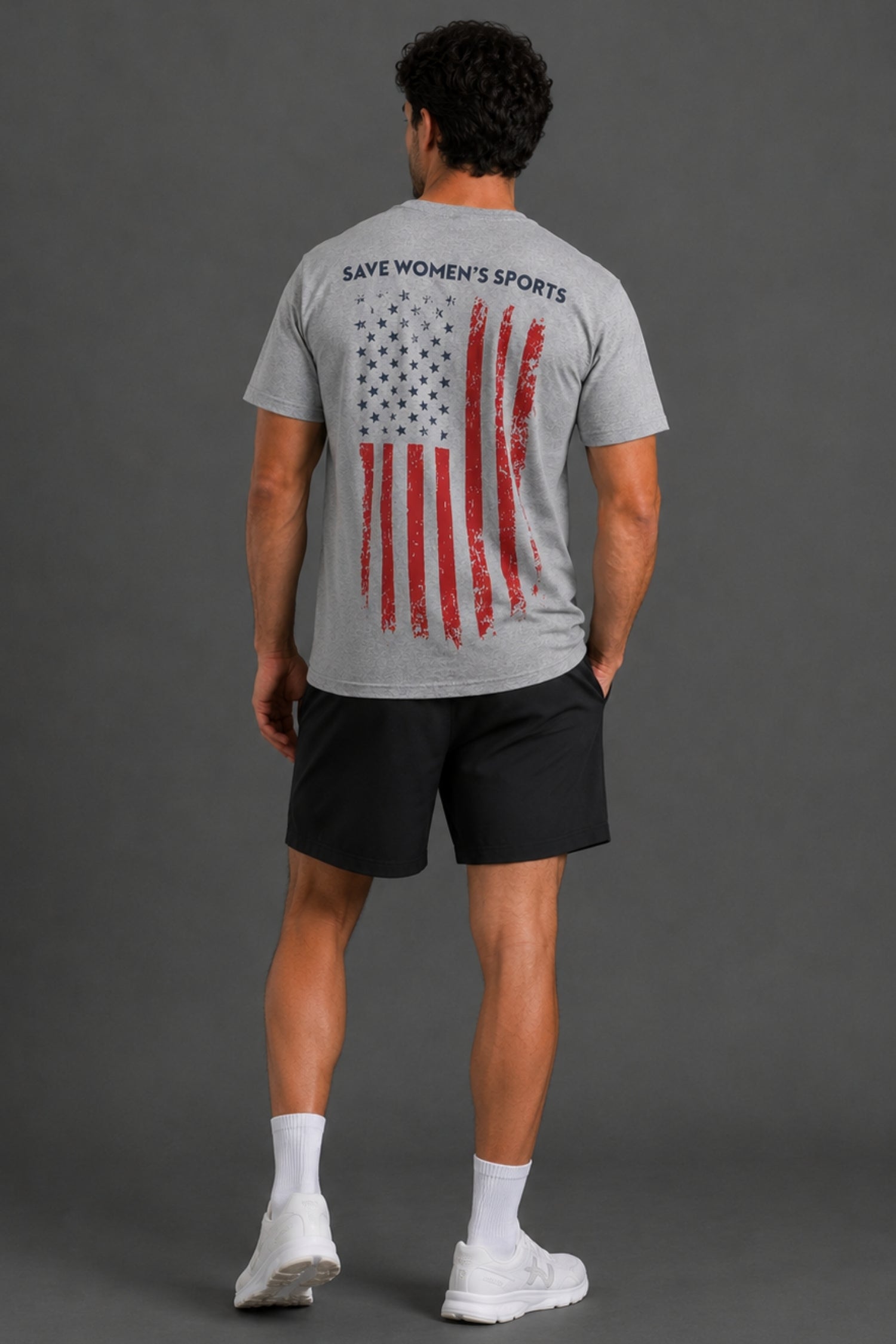 A man viewed from behind wearing a heather gray t-shirt with a distressed American flag graphic and the text 'SAVES WOMEN'S SPORTS' on the back, paired with black athletic shorts, white socks, and white sneakers against a dark gray studio background. || Heather Gray