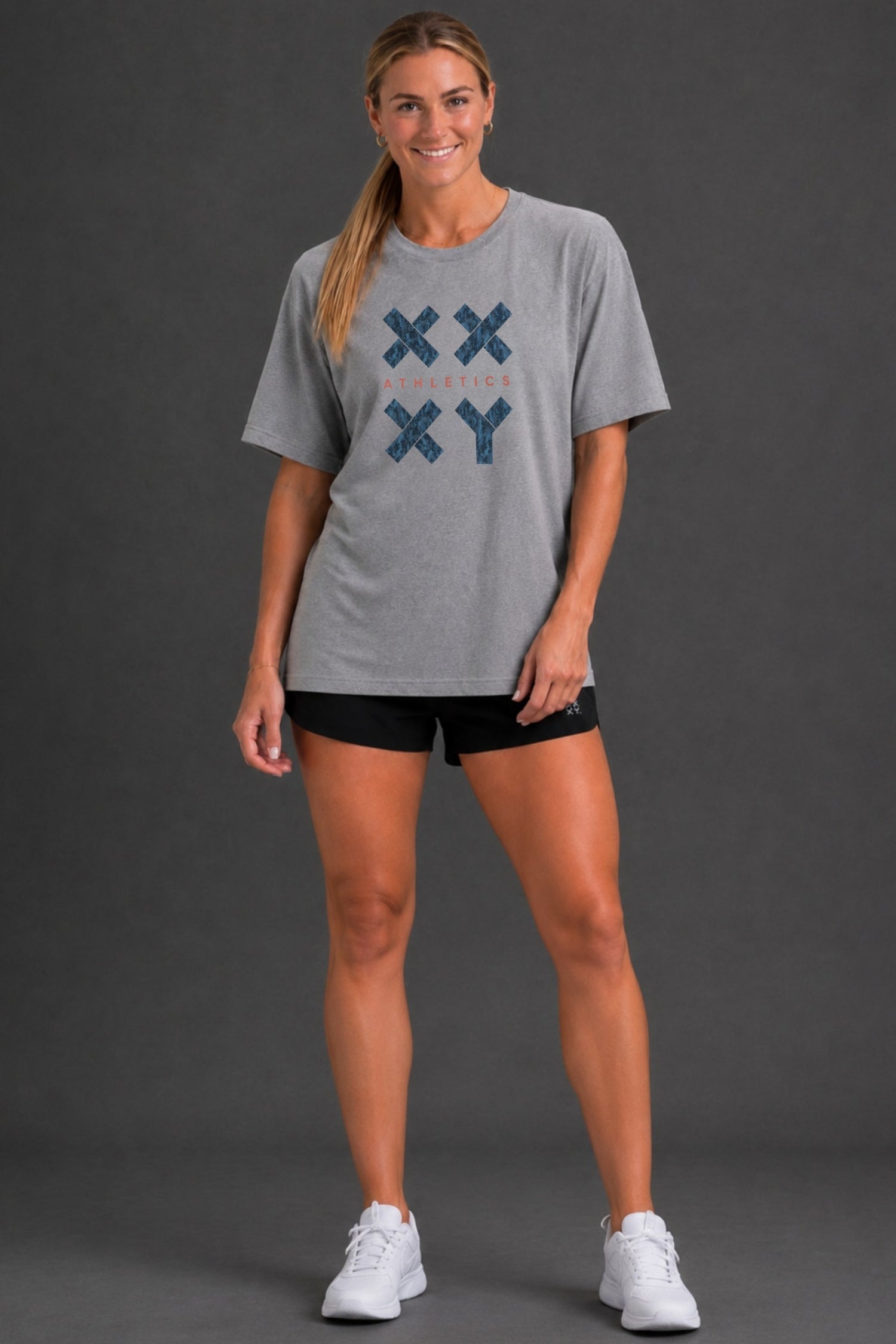 A woman modeling a heather gray short-sleeve t-shirt with a blue and orange graphic on the front, paired with black athletic shorts and white sneakers, against a dark gray studio background. || Heather Gray