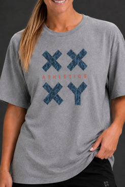 A woman wearing a heather gray short-sleeve t-shirt with a blue patterned 'XXYY' graphic and the word 'ATHLETICS' in coral text on the front, shown against a dark gray background. || Heather Gray