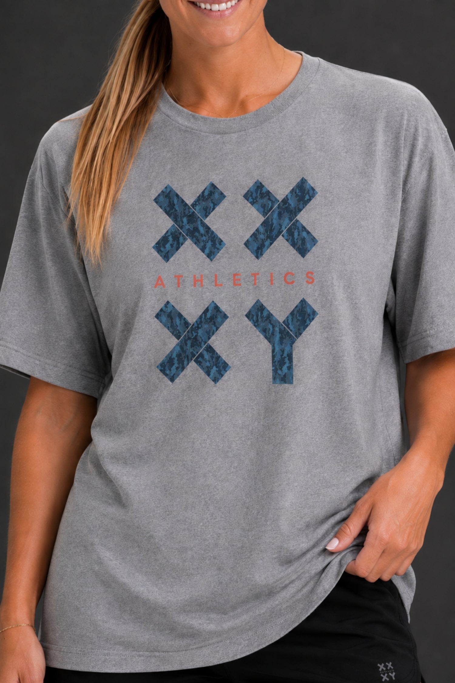 A woman wearing a heather gray short-sleeve t-shirt with a blue patterned 'XXYY' graphic and the word 'ATHLETICS' in coral text on the front, shown against a dark gray background. || Heather Gray