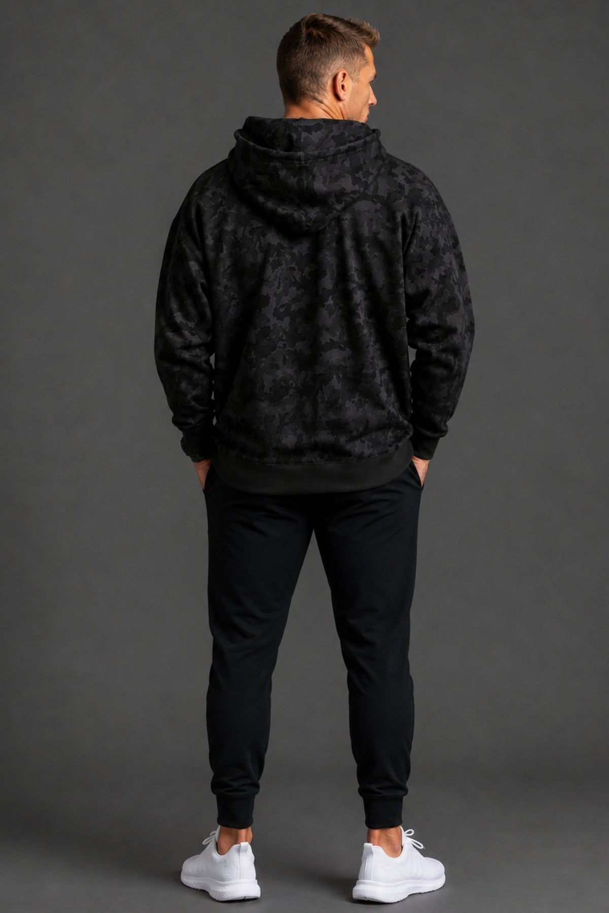 Male model shown from the back wearing a black camo hoodie, black joggers, and white sneakers against a dark gray studio background.