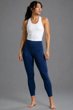 A woman modeling high-waisted, ankle-length leggings in a deep navy blue color, paired with a white tank top, against a solid gray studio background. || Navy