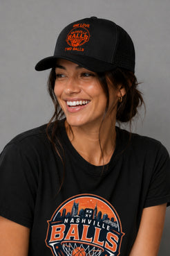 A woman smiling while wearing a black trucker-style baseball cap and a black graphic T-shirt. The cap and shirt both feature an orange and white circular 'Nashville Balls' design with a basketball theme. || Black