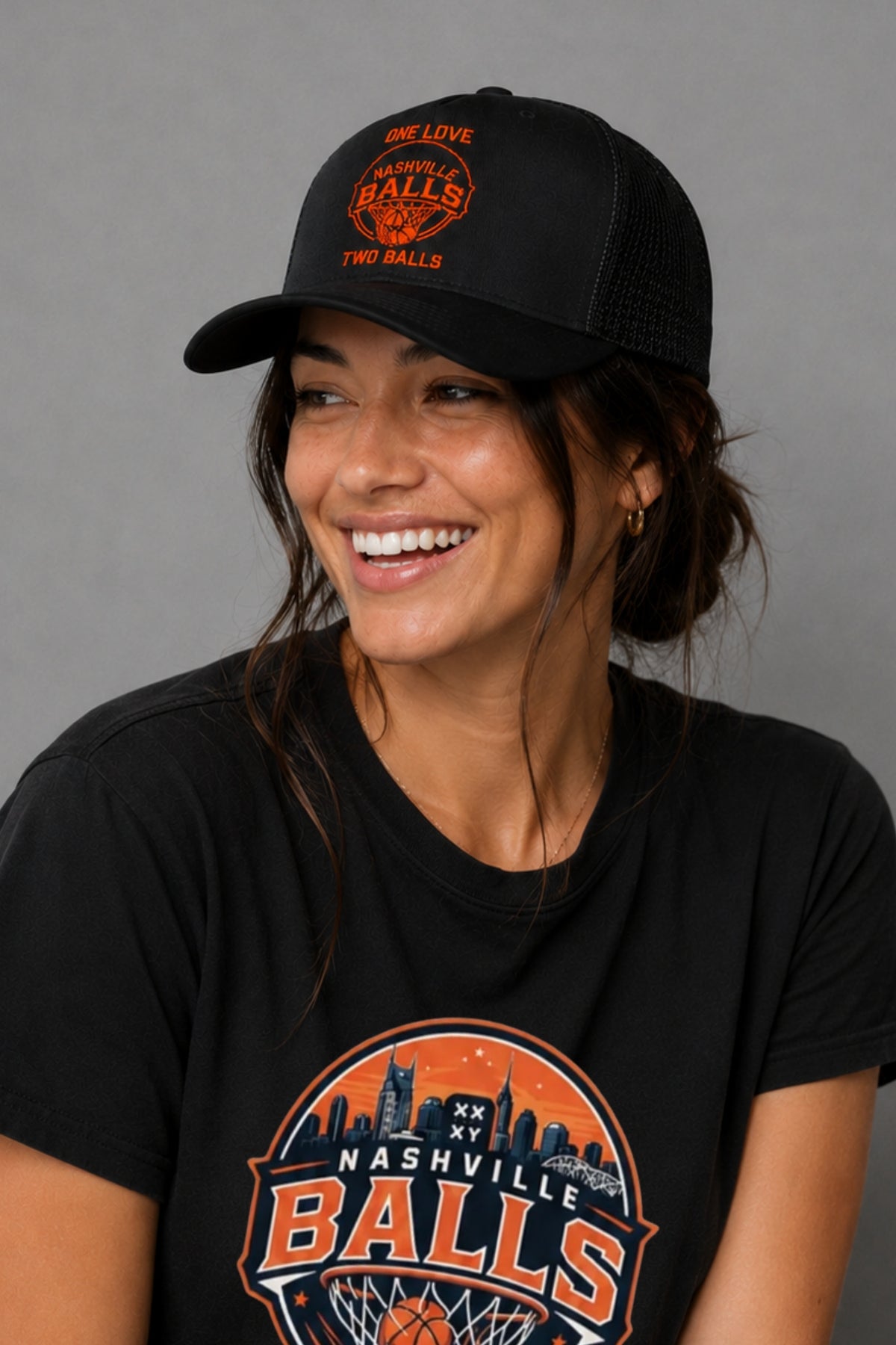 A woman smiling while wearing a black trucker-style baseball cap and a black graphic T-shirt. The cap and shirt both feature an orange and white circular 'Nashville Balls' design with a basketball theme. || Black