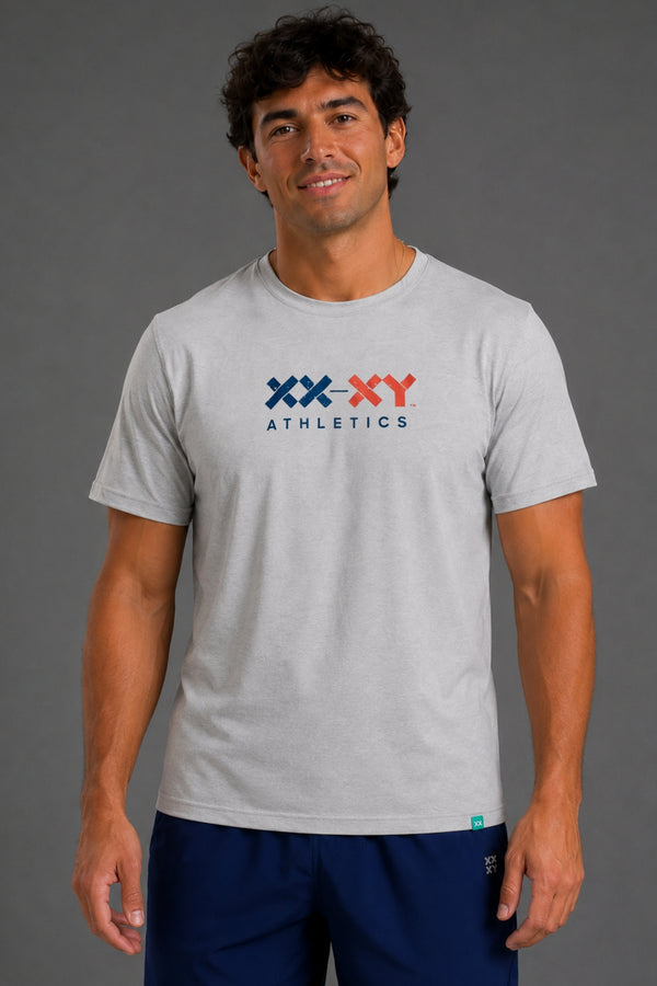 A man wearing a light gray heathered t-shirt with a navy and red 'XX-XY ATHLETICS' logo on the chest, paired with navy blue athletic shorts, against a solid gray studio background. || Light Heather Gray - 1