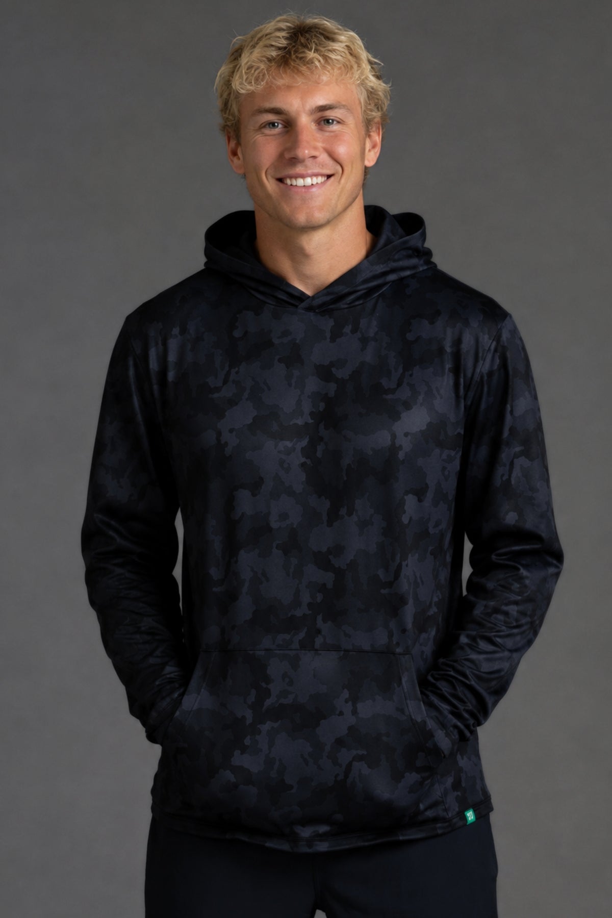 A smiling blonde man wearing a dark navy hooded sweatshirt with a subtle tonal abstract pattern, standing against a solid dark gray studio background.
