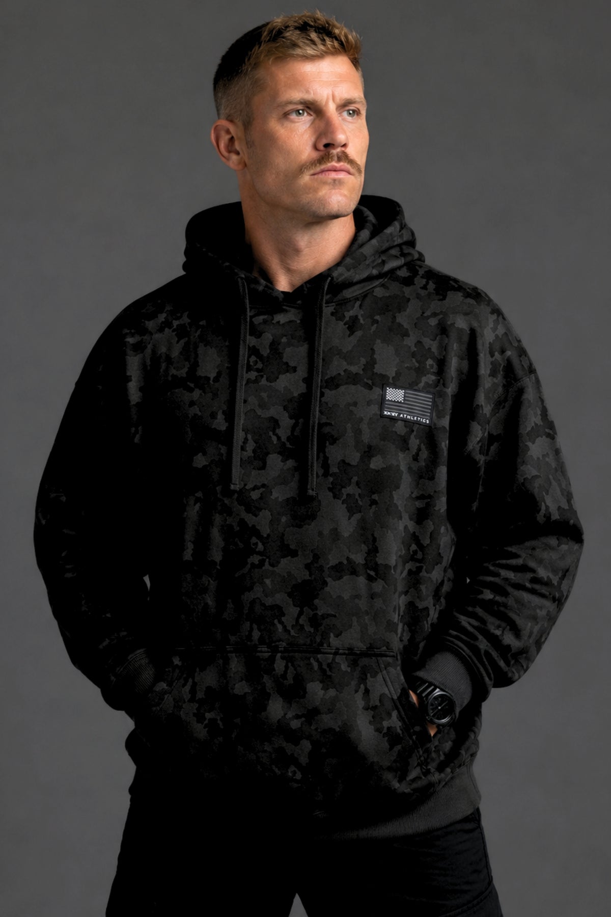 Male model shown from the front wearing a black camo hoodie, black joggers, and white sneakers against a dark gray studio background.