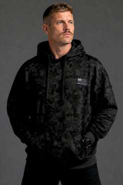 Male model shown from the front wearing a black camo hoodie, black joggers, and white sneakers against a dark gray studio background. || Black Camo