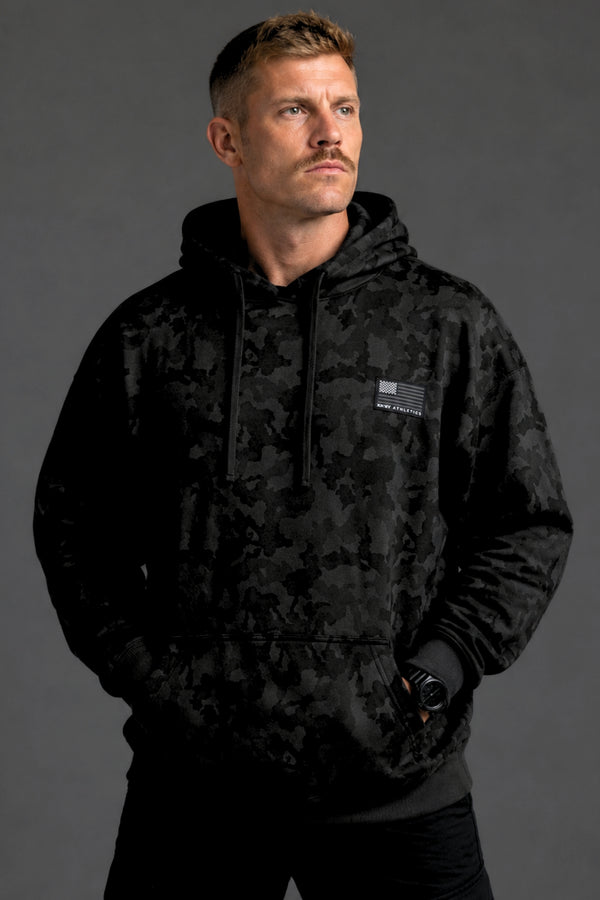 Male model shown from the front wearing a black camo hoodie, black joggers, and white sneakers against a dark gray studio background. || Black Camo